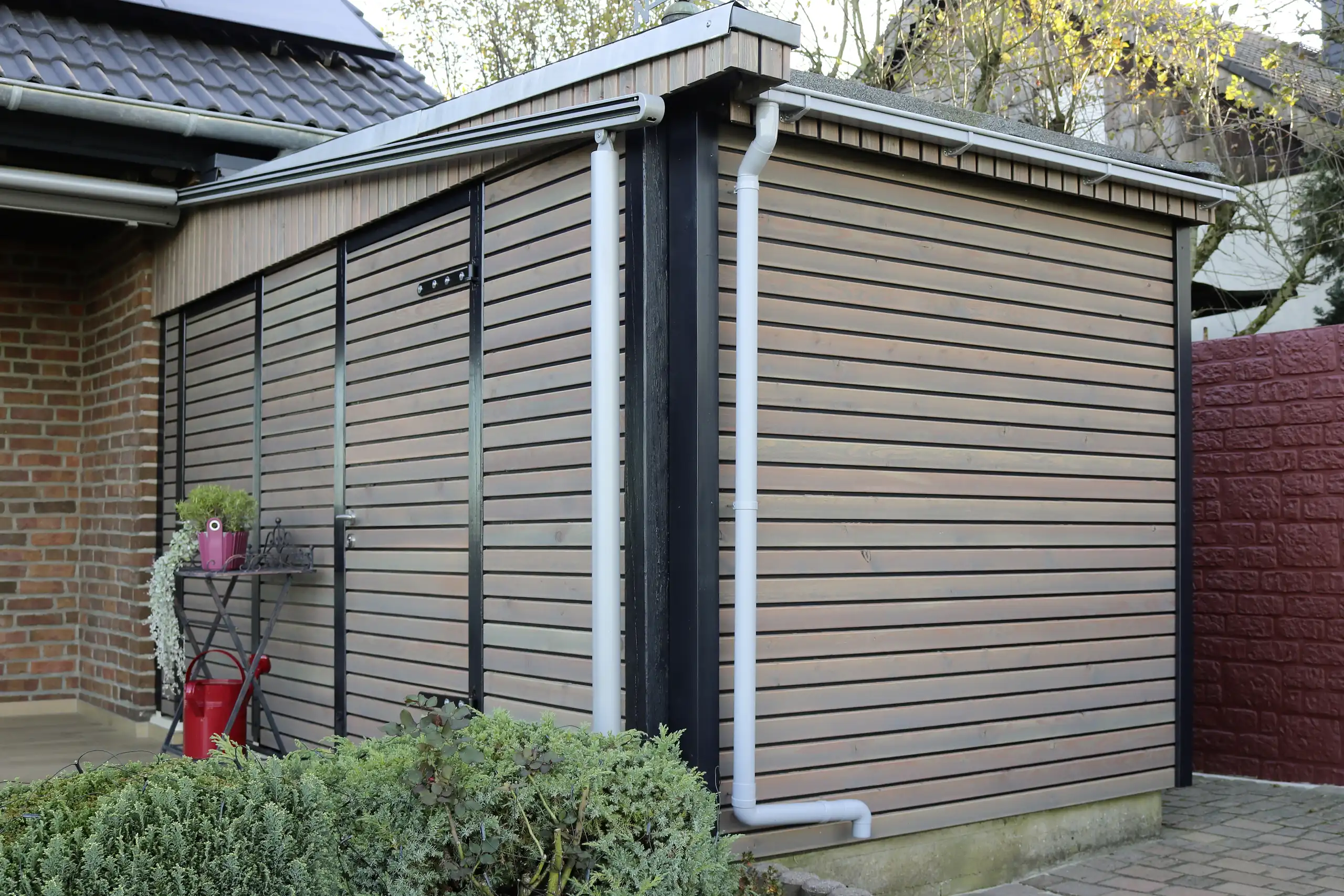 Gartenhütte mit grau lasierter Holzfassade, schwarzen Holzpfosten und weißem Regenfallrohr